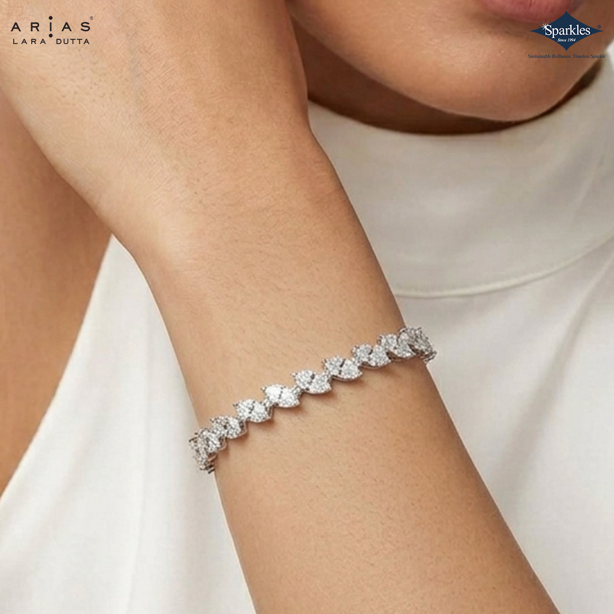 A close-up of a model's wrist wearing a Lara Dutta Arias bracelet, featuring a row of sparkling heart-shaped clear stones in a silver-toned setting. The model is wearing a white top, and the Lara Dutta brand logo is visible in the top left corner.