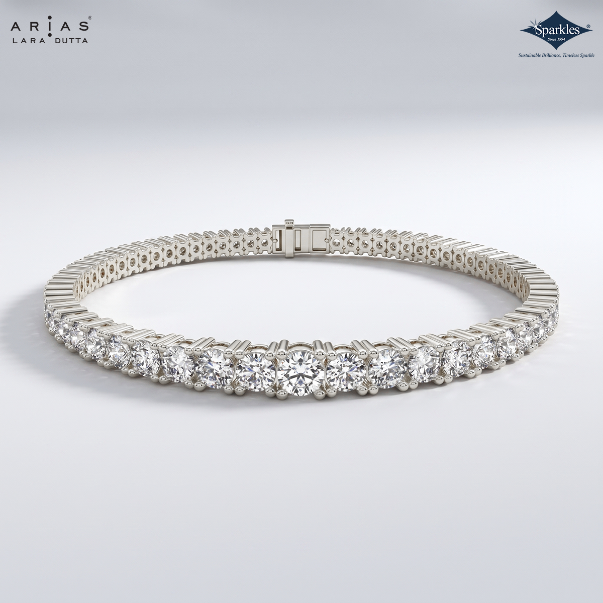 A silver tennis bracelet with a row of round-cut clear gemstones, displayed on a light gray gradient background. The ARIAS Lara Dutta brand logo is visible in the top left, and the Sparkles gem brand logo is in the top right.