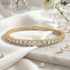 A gold tennis bracelet with clear round-cut gemstones, displayed on draped white fabric and a marble slab with pearl pins and white flowers in the background. Arias Lara Gutta branding is visible in the top left, and Sparkles jewelry branding in the top right.