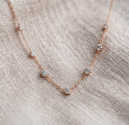 A delicate rose gold chain necklace with five small, clear round-cut gemstones spaced along the chain, displayed on a textured beige linen fabric.