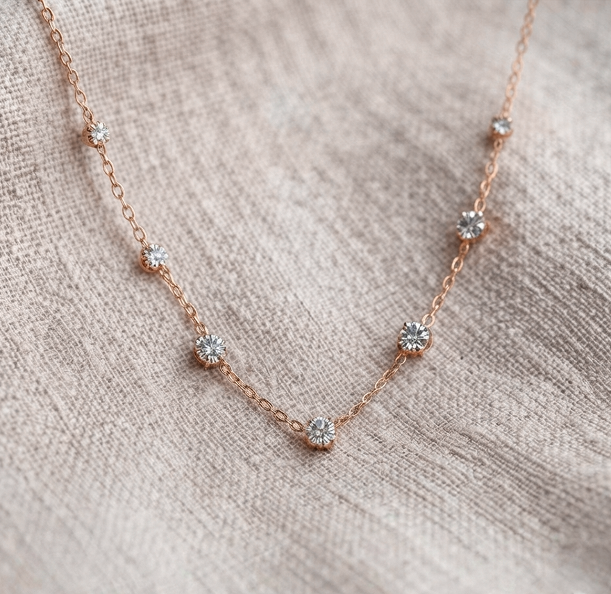 A delicate rose gold chain necklace with five small, clear round-cut gemstones spaced along the chain, displayed on a textured beige linen fabric.