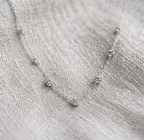 A delicate silver chain necklace with five small, clear flower-like cluster beads spaced along the chain, displayed on a textured beige linen fabric.