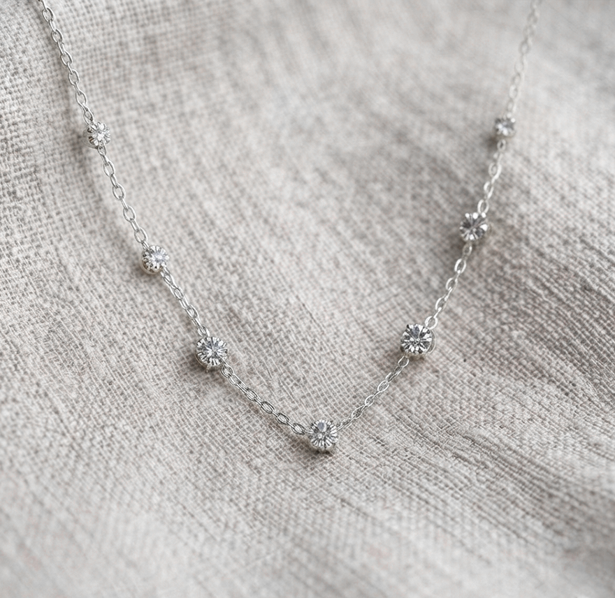 A delicate silver chain necklace with five small, clear flower-like cluster beads spaced along the chain, displayed on a textured beige linen fabric.