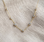 A delicate gold chain necklace with five small, clear round-cut gemstones spaced along the chain, displayed on a textured beige linen fabric background.