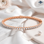 A rose gold tennis bracelet with a row of clear round-cut gemstones, displayed on a white marble block with white satin fabric, rose petals, and a pearl. The ARIAS LARA DUTTA logo is in the top left, and the Sparkle jewelry brand logo is in the top right.