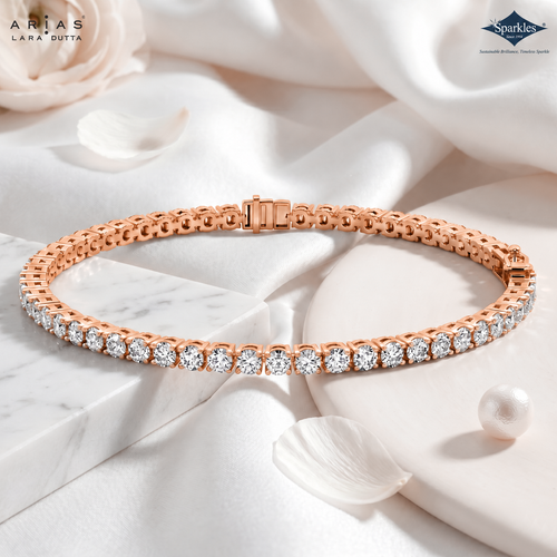 A rose gold tennis bracelet with a row of clear round-cut gemstones, displayed on a white marble block with white satin fabric, rose petals, and a pearl. The ARIAS LARA DUTTA logo is in the top left, and the Sparkle jewelry brand logo is in the top right.