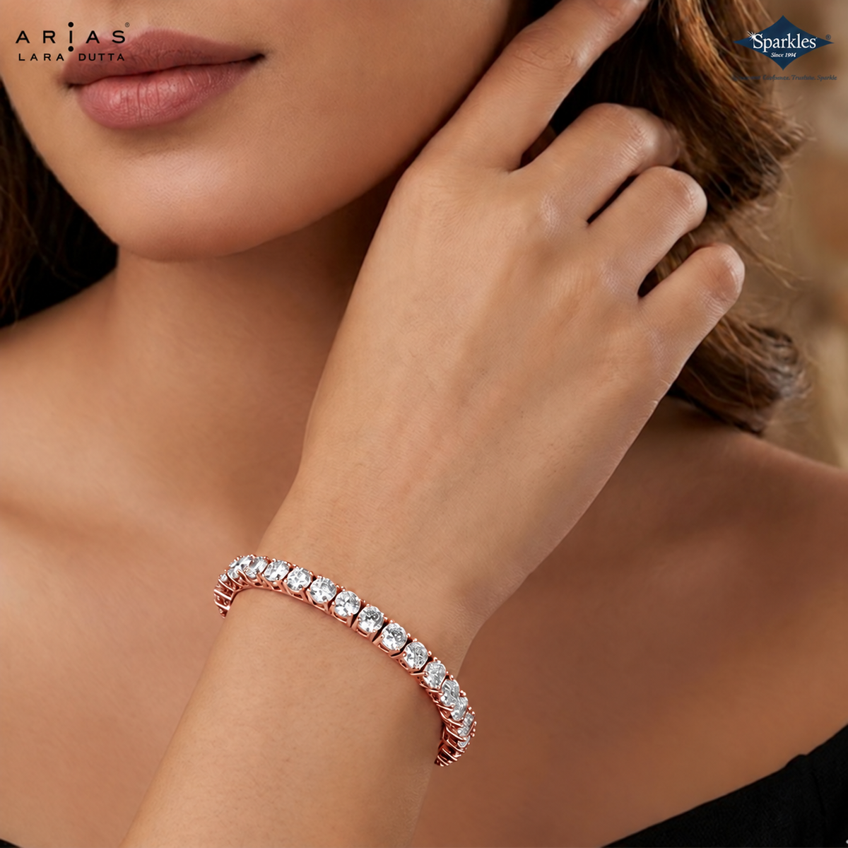 A gold tennis bracelet with clear round-cut gemstones worn on a woman's wrist, shown in a close-up lifestyle shot. The brand logos 'Arias Lara Ditta' and 'Sparkles' are visible in the corners.