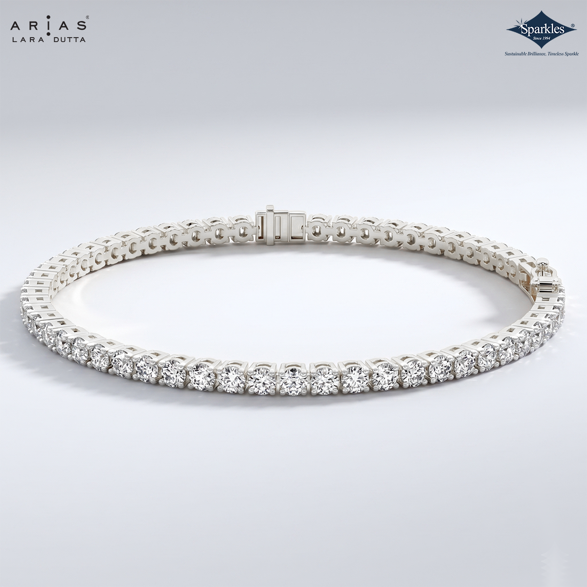 A silver tennis bracelet with round-cut clear stones, displayed in a circular shape on a light gray gradient background. The brand logos 'ARIA'S LAURA DUTTA' and 'Sparkles' are visible in the top corners.