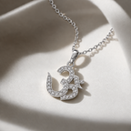 A silver-toned Om pendant necklace adorned with small clear stones, resting on a draped cream satin fabric.