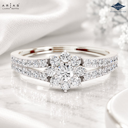A silver-tone diamond ring set by Arijas Lara Dutta, featuring a central floral cluster of round diamonds flanked by pavé-set diamond bands, displayed on a reflective white marble surface with soft white fabric in the background.