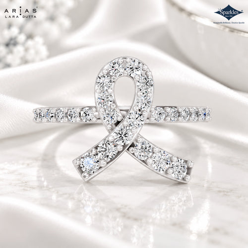 A diamond ring shaped like a ribbon, featuring a silver-toned metal base set with numerous clear round-cut diamonds. The ring is displayed on a white marble surface with draped white satin fabric in the background.