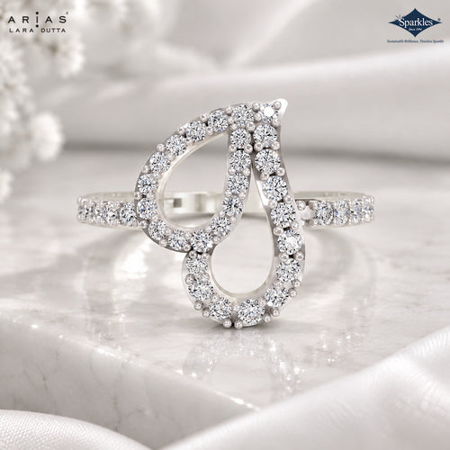 A silver-tone ring with two interlocking teardrop-shaped clusters of clear pavé-set stones, displayed on a white marble slab with white fabric in the background. The Arias Lara Dutta brand logo is visible in the top left, and the Sparkles Jewellery brand logo is visible in the top right.