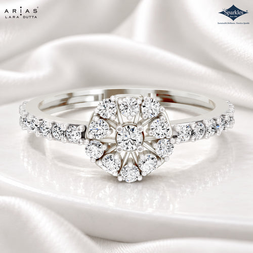 A silver-tone halo ring with a central round diamond and surrounding cluster diamonds on the band, displayed on white satin fabric. Arias Lara Dutta branding appears in the top left, and a Sparkle certification mark appears in the top right.