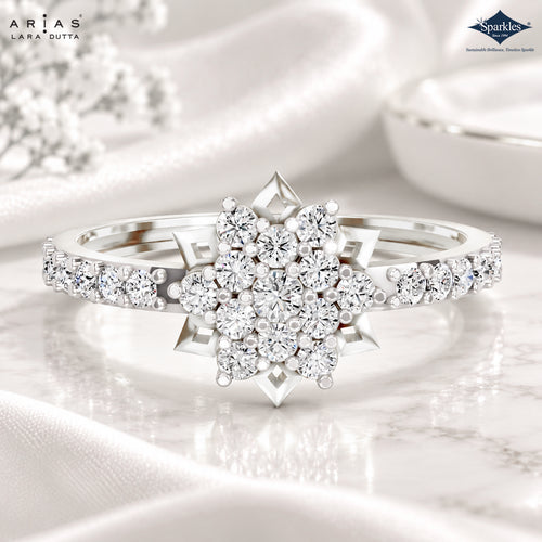 A silver-tone diamond ring with a starburst cluster of round diamonds on the center and smaller diamonds along the band, displayed on a white marble surface with draped satin fabric. The brand logos for Arias Lara Dutta and Sparkle are visible in the corners.