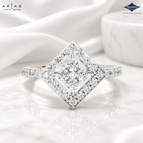 A diamond ring with a square-shaped pavé center and a diamond-cut halo setting, shown on a white marble surface with draped white fabric and a marble bowl in the background. The brand logos 'ARIA'S LARA DUTTA' and 'Sparkle' are visible in the corners.
