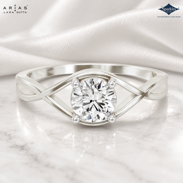 A silver-tone diamond ring with a round-cut center stone and an open, twisted band design, displayed on a white marble surface with draped satin in the background. The Arias Lara Dutta brand logo is visible in the top left, and the Sparkle ring maker logo is in the top right.
