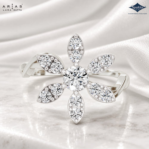 A silver-tone flower-shaped ring with multiple clear round and marquise-cut gemstones, displayed on a soft white satin fabric background. The brand logos 'Arias Lara Dutta' and 'Sparkles' are visible in the top corners.
