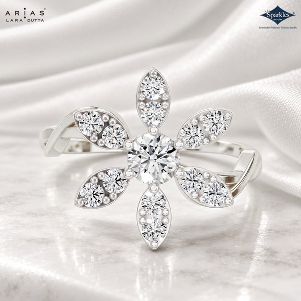 A silver-tone flower-shaped ring with multiple clear round and marquise-cut gemstones, displayed on a soft white satin fabric background. The brand logos 'Arias Lara Dutta' and 'Sparkles' are visible in the top corners.