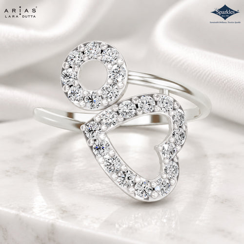 A silver-tone ring with a heart-shaped outline paved with clear stones and a circular oval accent, shown on a soft white satin background. ARIA'S LARA DUTTA branding appears in the top left, and the Sparkle logo appears in the top right.