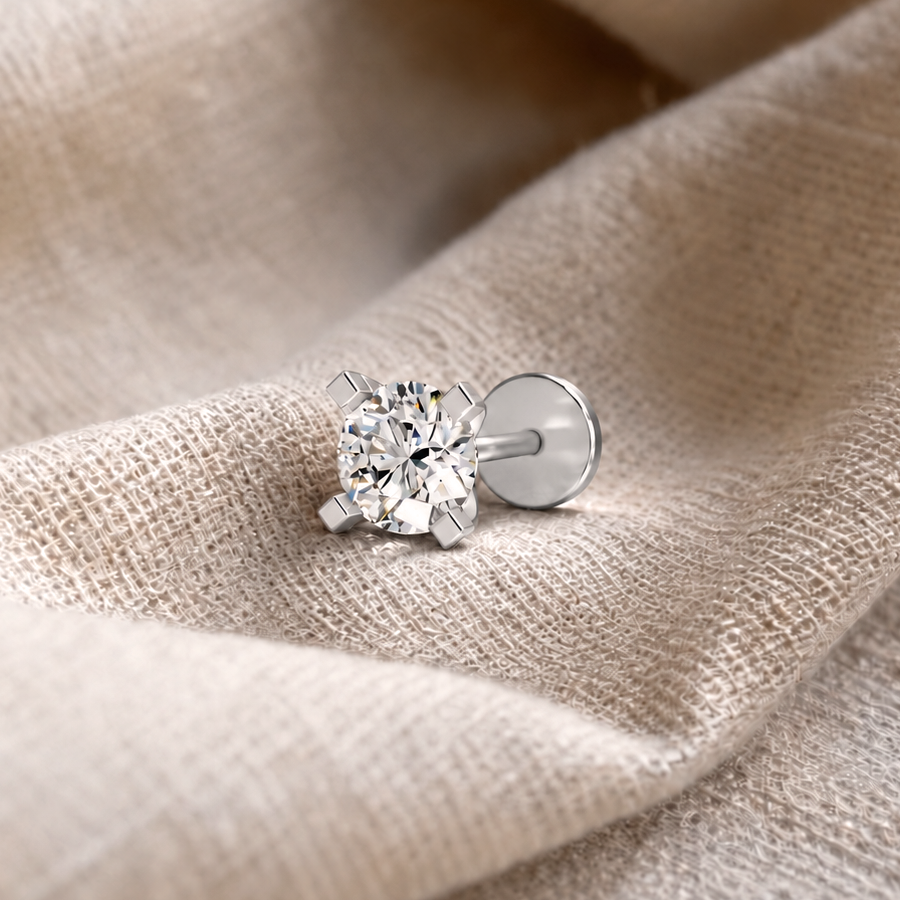 A single solitaire stud earring with a round brilliant-cut clear gemstone in a four-prong silver setting, displayed on a textured beige fabric background.