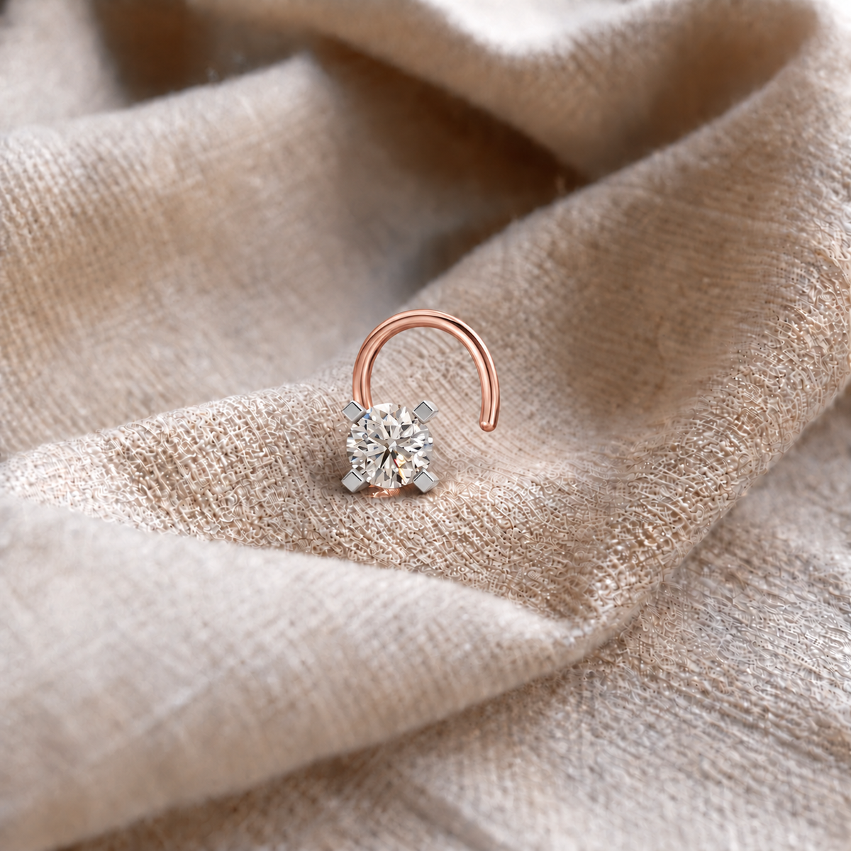 A close-up of a small curved rose gold ring with a clear solitaire gemstone in a four-prong setting, resting on draped beige fabric.