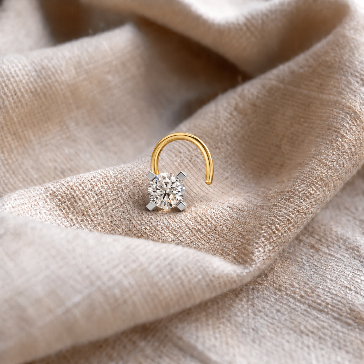 A gold hoop nose ring with a single round-cut clear gemstone in a silver-toned setting, resting on draped beige linen fabric.