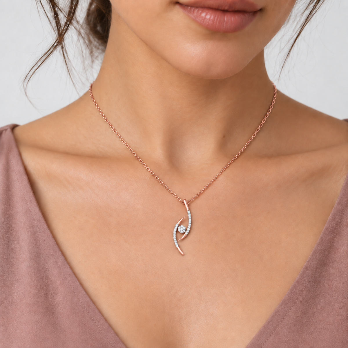 A close-up of a woman wearing a delicate rose gold necklace with a curved pendant set with small clear stones, paired with a mauve top against a light gray background.