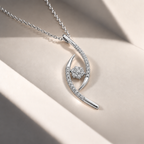 A silver pendant necklace with a curved, openwork design set with small clear stones and a central round stone cluster, displayed on a beige surface with soft shadows.