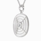 A silver oval pendant necklace featuring concentric circular patterns, a textured outer edge, and a small central clear gemstone, suspended from a silver chain against a light gray background.