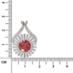 A teardrop-shaped pendant with a round red center stone and surrounding silver-toned rays set with small clear stones, shown next to a centimeter ruler for scale on a white background.