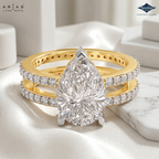 A luxury wedding ring set by Aria's Lara Dutta, featuring a pear-cut diamond center stone on a yellow gold band with pavé-set diamonds, displayed on a white satin fabric with marble accents.