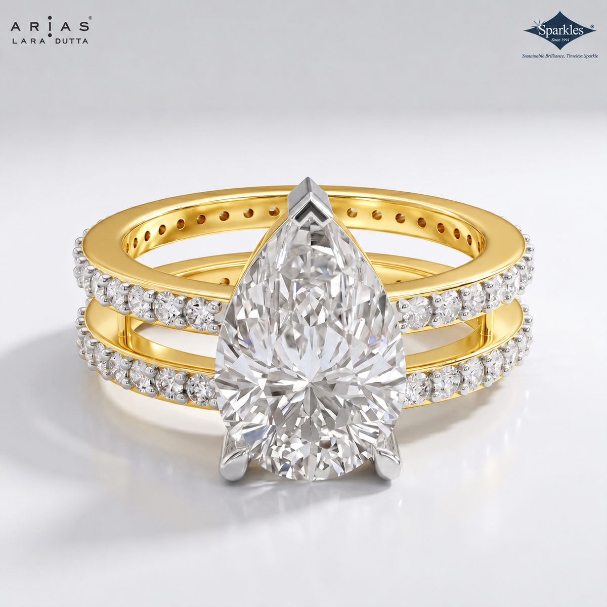 A double-band ring set by ARIAS LANA DUTTA, featuring a large pear-cut center stone in a silver-toned setting, with gold bands accented by pavé diamonds. The Sparkles hallmark logo is visible in the top right corner.