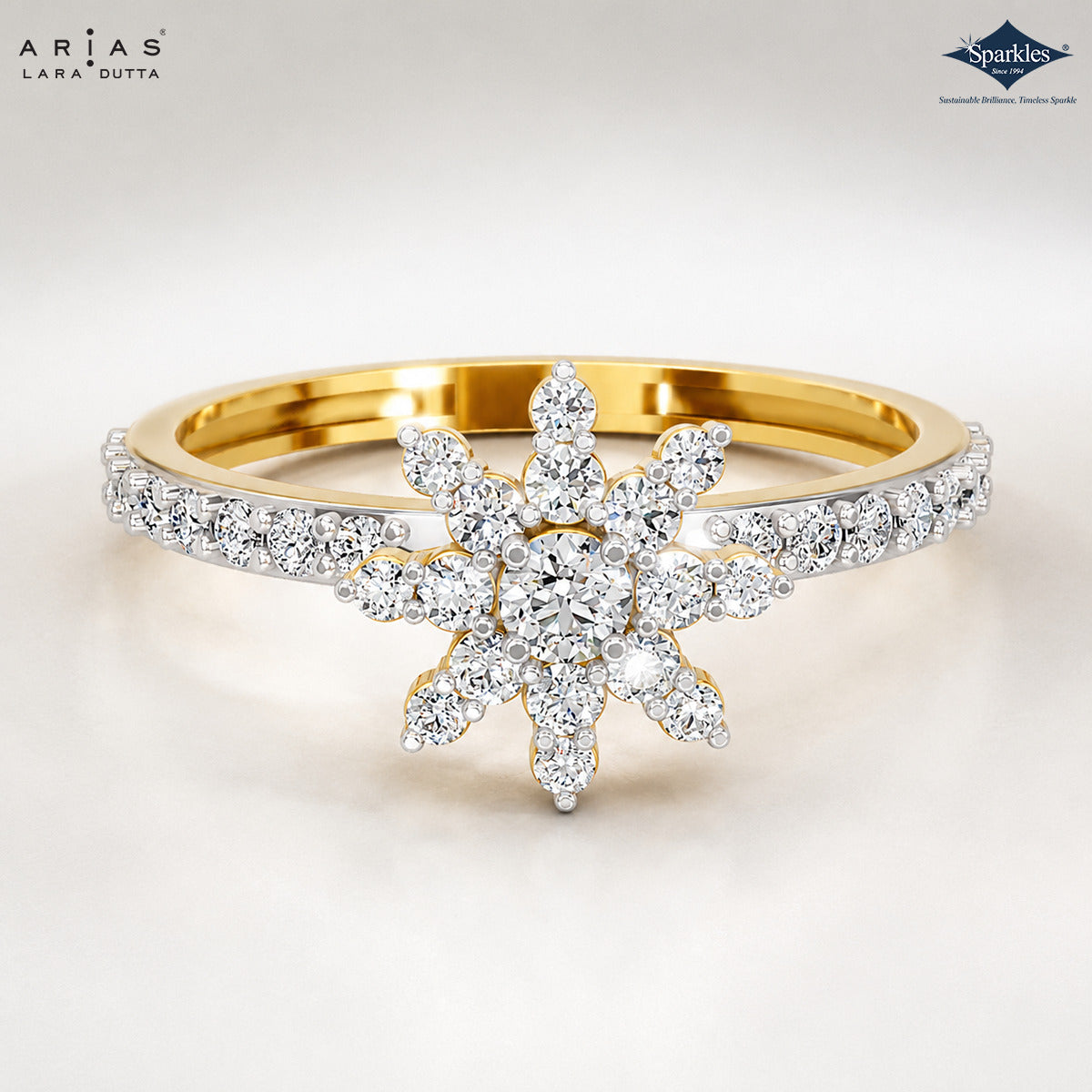 A gold and white gold ring featuring a central snowflake-like cluster of round brilliant-cut diamonds, with a pavé-set diamond band, shown on a clean white background. ARIA DUTTA branding is visible in the top left, and Sparkles jewelry branding is in the top right.